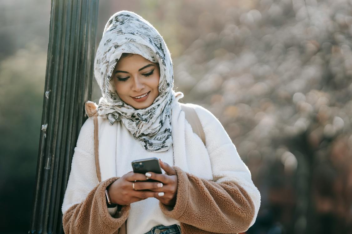 Free Cheerful young Muslim lady wearing warm coat and floral hijab leaning on pillar while surfing smartphone outdoors Stock Photo