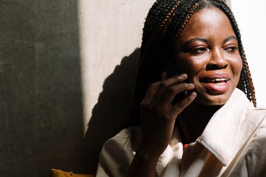 Free Woman with braids smiling while talking on her mobile phone indoors. Sunlit portrait. Stock Photo