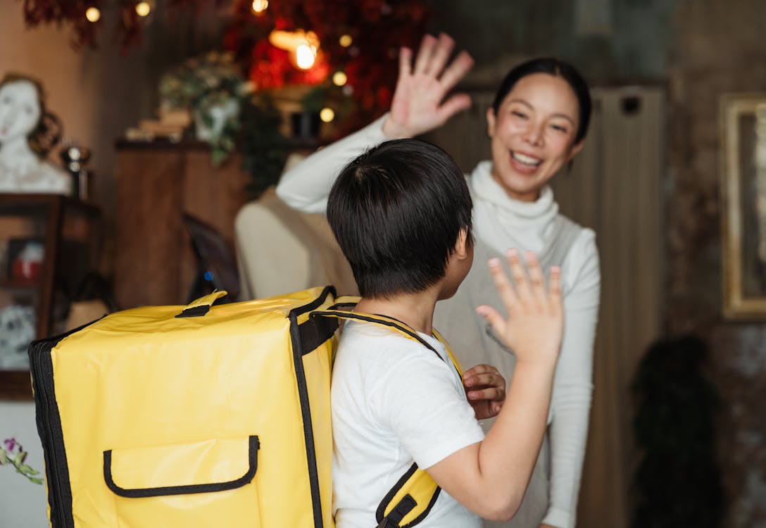 Free Happy ethnic female cafe worker in apron wishing farewell to boy with food delivery backpack helping in family business Stock Photo
